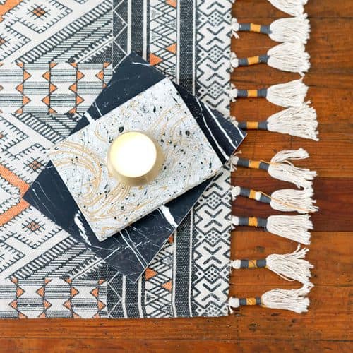 A lit candle sits on a decorative marbled tray, with two black and white patterned books underneath—perfect inspiration for exploring different types of self care—placed on a geometric patterned rug with white tassels by a wooden surface.