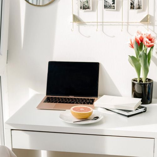 A clean white desk with a laptop, a plate holding half a grapefruit, stacked notebooks, and a vase of pink tulips. Sunlight streams in, creating a bright workspace that inspires your daily self-care routine.