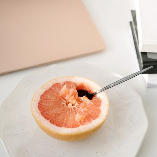 A halved grapefruit with a spoon inside rests on a white plate, hinting at a mindful daily self-care routine. Nearby, a closed laptop and a stack of books are partially visible on the white surface.