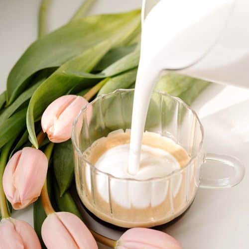 A glass cup of coffee is being topped with steamed milk, surrounded by soft pink tulips and green leaves on a light surface—perfect for inspiring morning self-care ideas.