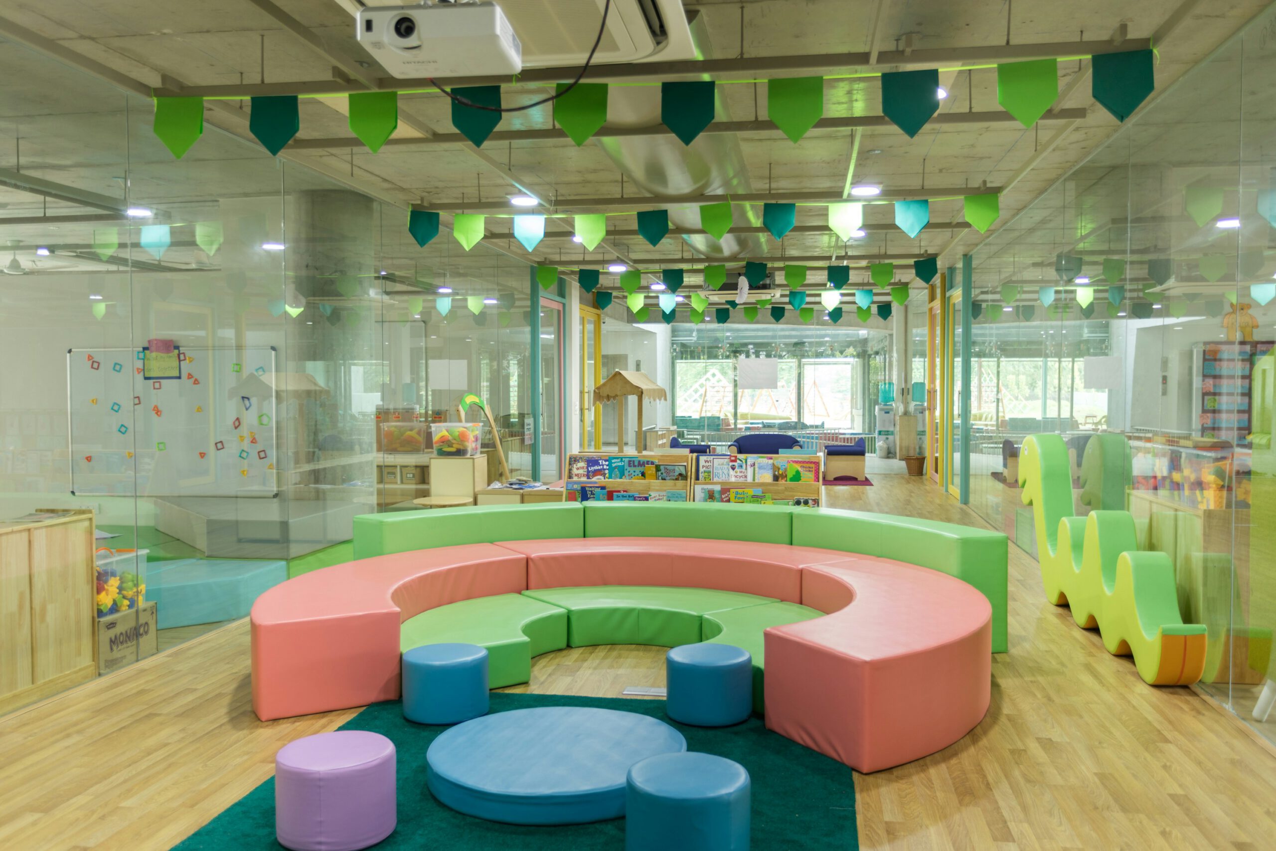 Farbenfroher, moderner Kindergartenraum mit runden Sitzmöbeln, Bücherregal und Spielbereichen, mit Glaswänden und Holzfußboden.
