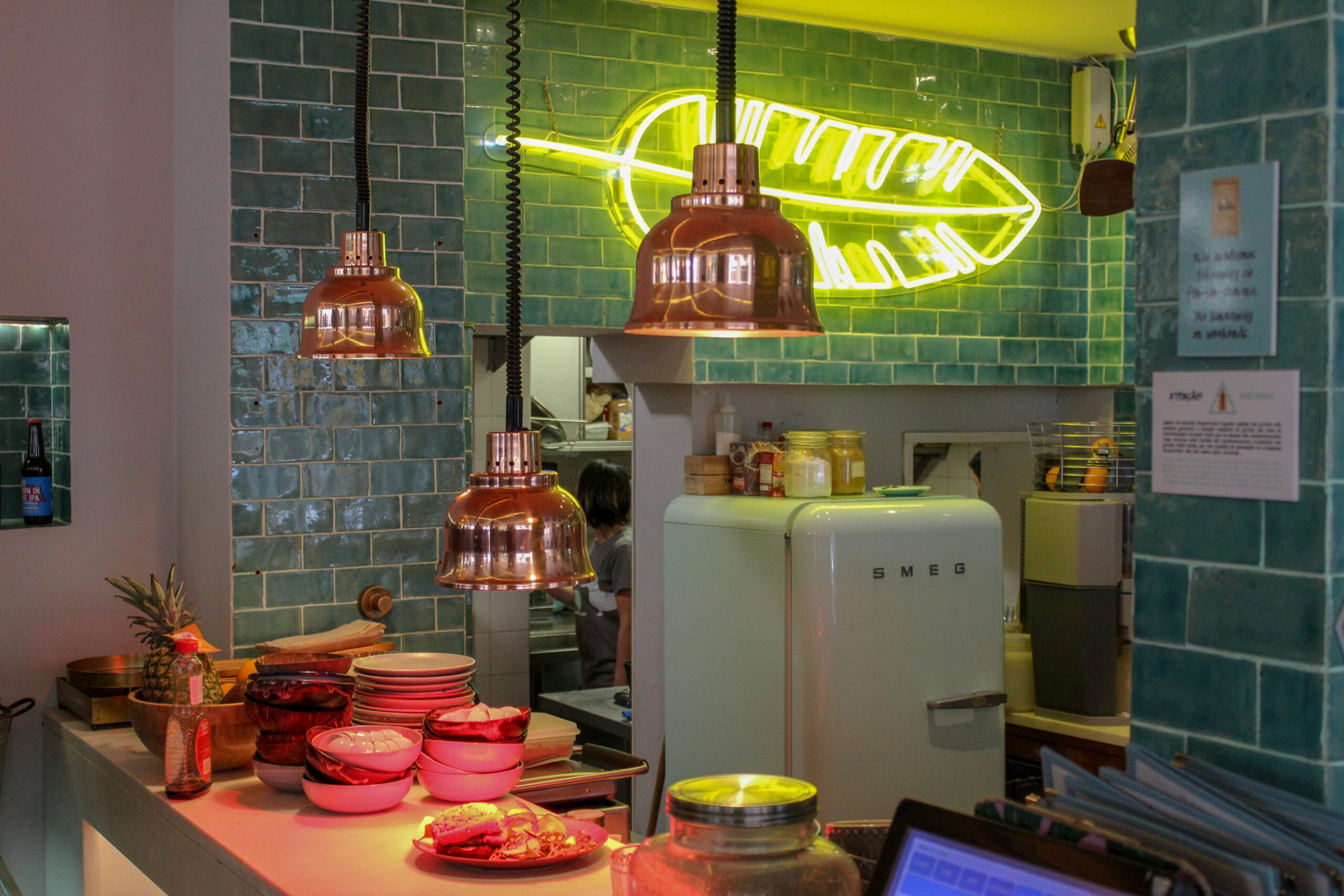 Stilvolle Gastronomie-Theke mit Retro-Kühlschrank, Kupferlampen, türkisen Fliesen und Neon-Leuchtschild in Federform.