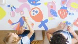 Zwei Kinder basteln konzentriert an einem Tisch mit bunten Papierfiguren. Die ruhige, fokussierte Stimmung lässt auf eine gute Raumakustik in der Kita schließen, die das kreative Arbeiten unterstützt und Lärm reduziert.