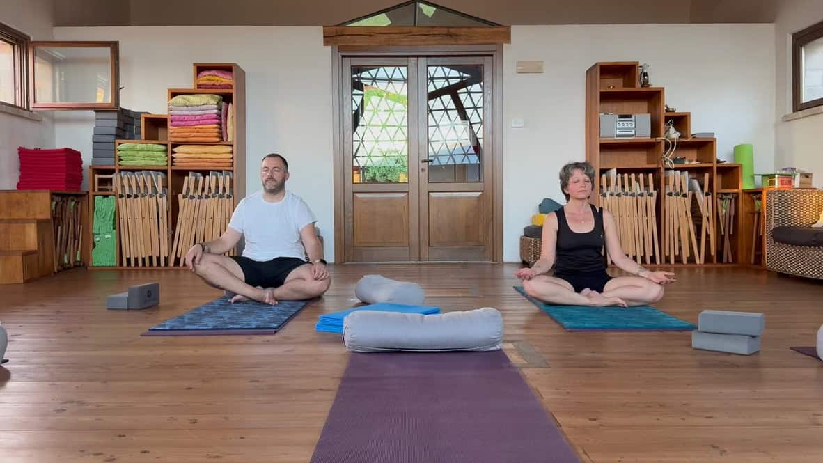 Due persone praticano yoga sedute in sala con attrezzi e tappetini.