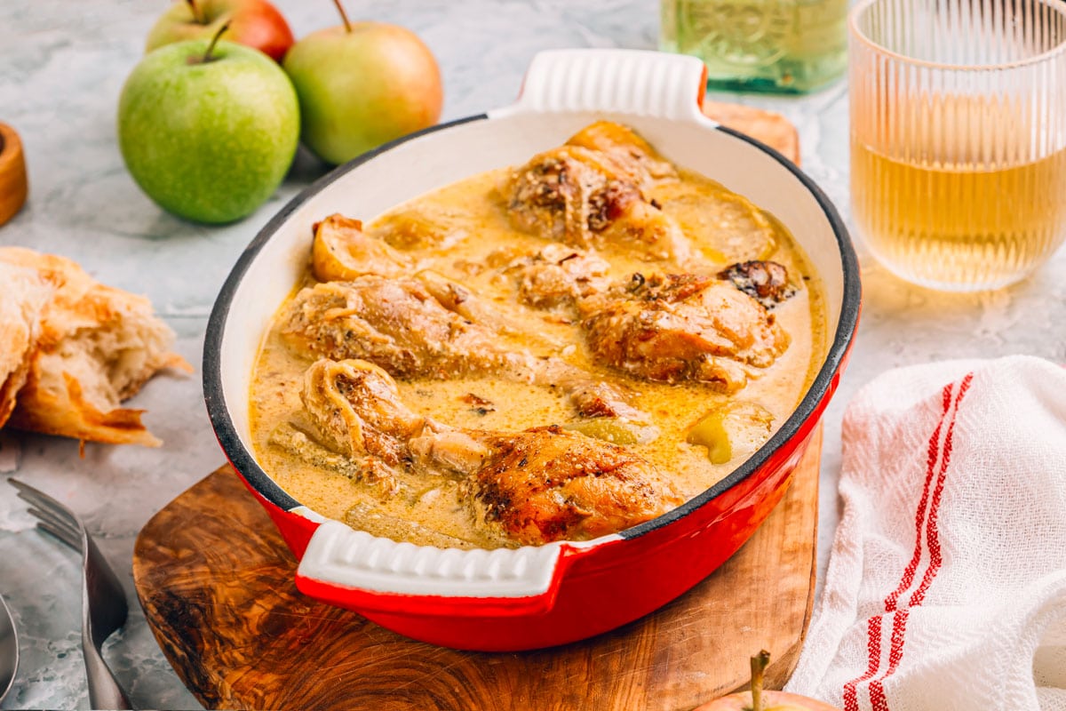 shutterstock 2393687359 ©julia mikhaylova poulet de normandie servi dans moule en fonte recouvert demail pommes verre de cidre sur fond gris marbre. cuisine francaise. lidee de recette dautomne