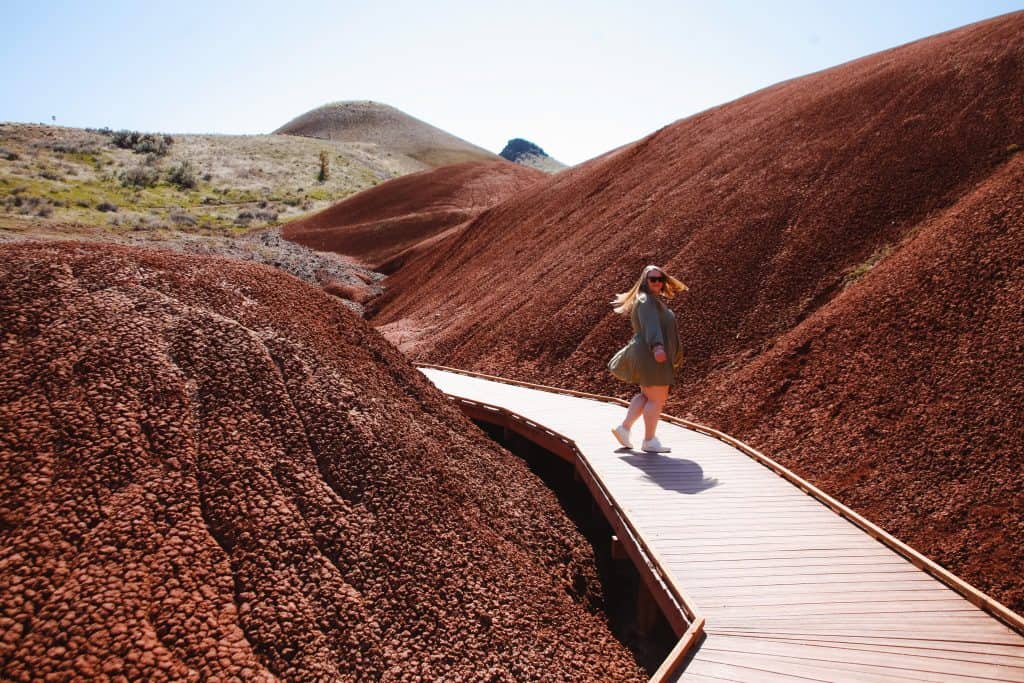 Michele in a casual olive dress and sunglasses walks along a curving wooden boardwalk amidst the striking red and brown volcanic hills of John Day Fossil Beds National Monument, Oregon, under a clear blue sky.