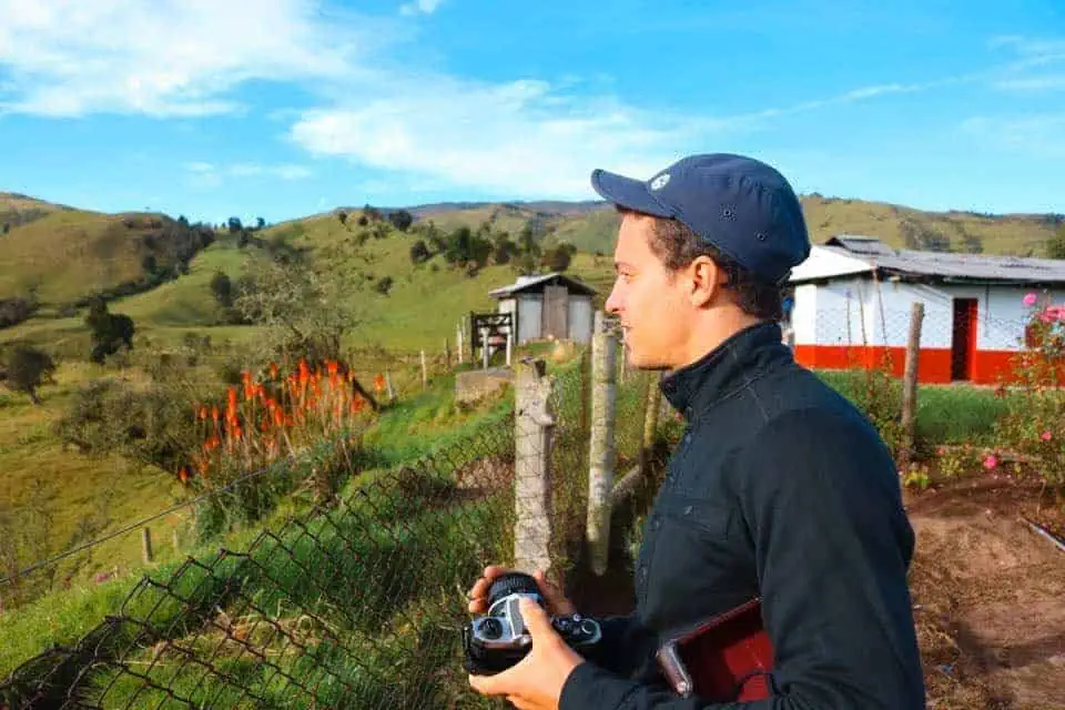 A man stands at a wooden fence holding a camera and looking out among the valley.
