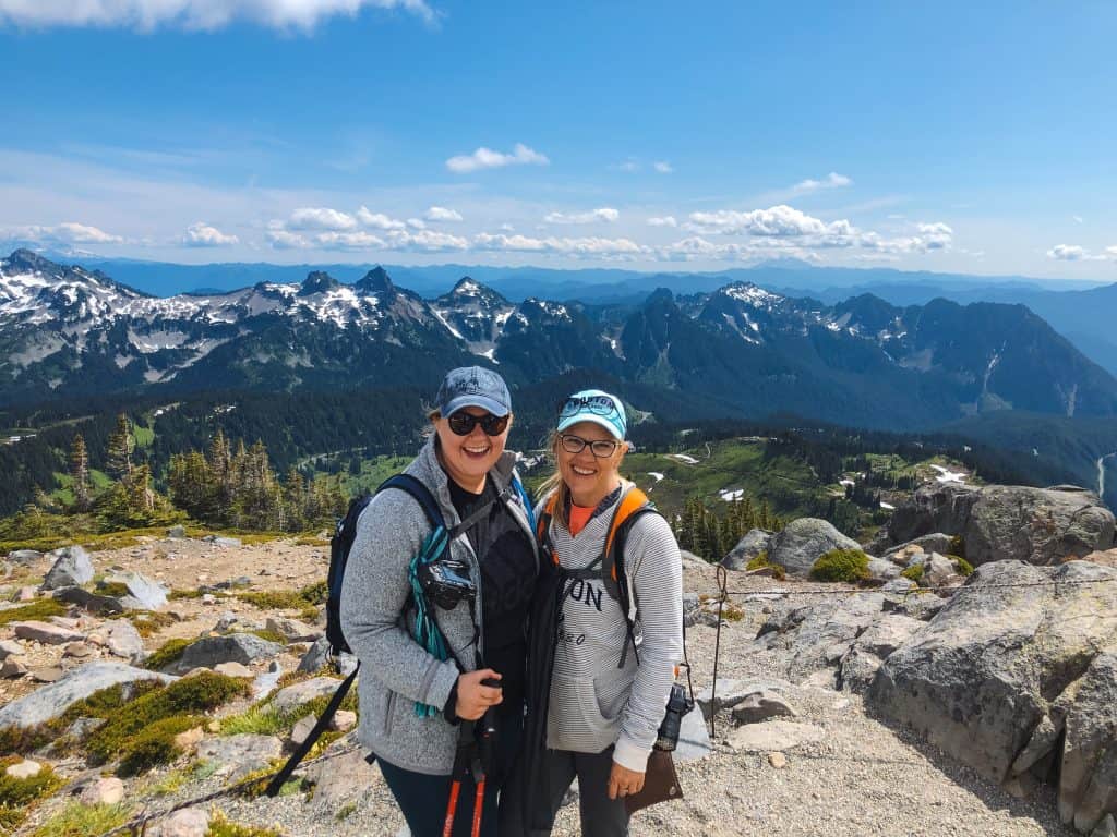 Two hikers are smiling for a photo on Mount Rainier in Washington, with stunning panoramic views of jagged peaks and lush valleys in the background. They're equipped with backpacks, sunglasses, and trekking poles, ready for adventure amidst the breathtaking landscape.