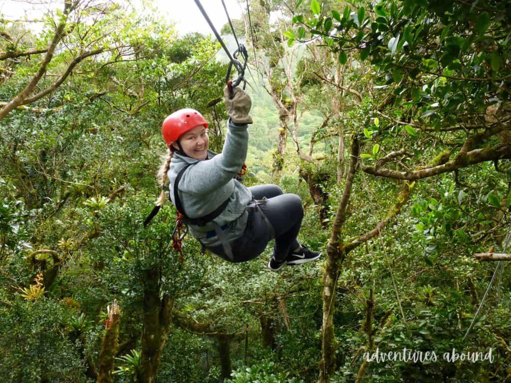 Michele wearing a red helmet and harness is ziplining with a delighted expression among dense green foliage in Costa Rica.