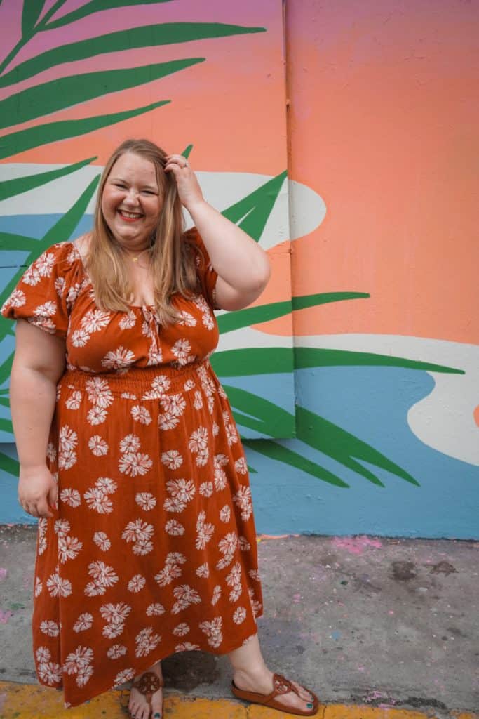 Michele, a woman with blonde hair, in a vibrant orange floral dress is smiling and playfully touching her hair, standing against a colorful mural with large green palm leaf patterns. She's wearing brown sandals and appears to be enjoying herself on a sunny day on vacation.