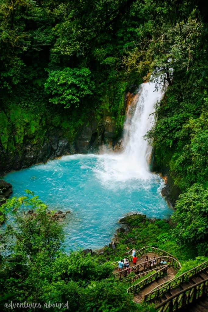 A vibrant scene at Rio Celeste in Costa Rica, featuring the famous turquoise water plunging into a pool from a waterfall surrounded by lush greenery. Visitors can be seen on a viewing platform at the bottom, taking in the natural beauty of the area.