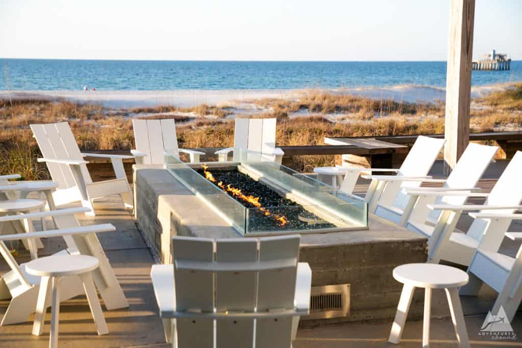 The image shows an outdoor fire pit area at a Gulf Shores hotel, featuring white Adirondack-style chairs arranged in a circle around a modern, rectangular fire pit with glass sides. The fire is lit, with flames dancing over black stones. In the background, the beach and ocean are visible, with dune grasses leading down to the shore. A wooden pier can be seen in the distance.