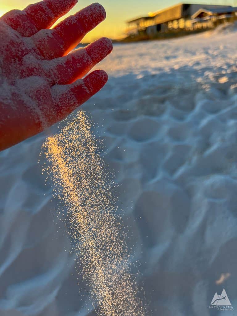 a close-up of a hand letting fine, white sand trickle through its fingers at sunset. The golden light highlights the grains of sand against the soft, rippled surface of the beach. The Lodge hotel in the background is slightly out of focus but glowing bright in the golden hour