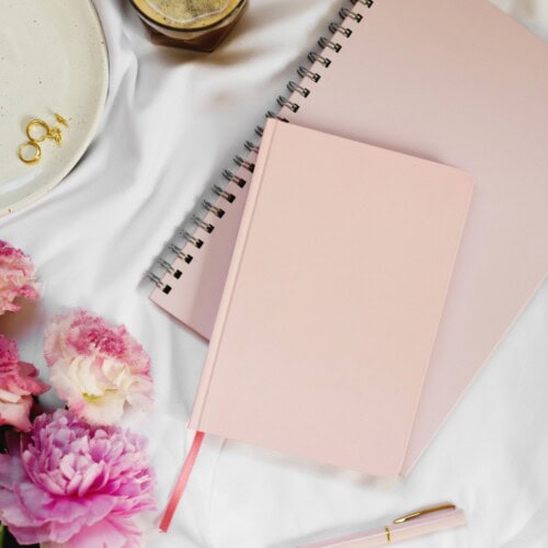365 Journal Prompts For a Year 4 A flat lay of a pink notebook, a spiral-bound journal with journal prompts for a year, pink pen, a plate with gold rings, a cup of coffee, and pink flowers on a white surface.