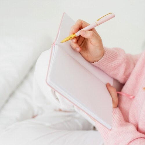 365 Journal Prompts For a Year 5 A person in a pink sweater sits on a white bed, holding an open blank notebook and a pink pen, ready to write with 365 journal prompts for a year.