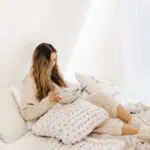 A woman with long hair sits comfortably on a bed with white pillows and a chunky knit blanket, exploring journaling prompts for mental health in a bright, cozy room.