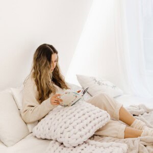 A woman with long hair sits comfortably on a bed with white pillows and a chunky knit blanket, exploring journaling prompts for mental health in a bright, cozy room.