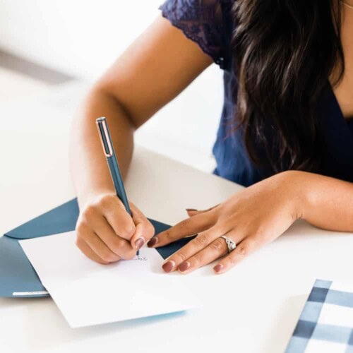 A person with long dark hair writes self confidence journal prompts on a blank white card with a pen, next to a blue envelope on a white table.