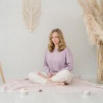 A woman with blonde hair sits cross-legged on a light pink blanket, meditating with eyes closed. Surrounded by candles, pampas grass, and boho decor, she enjoys a calm room as she reflects on mindfulness journal prompts.