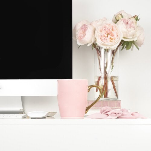 A white desk with a computer monitor, a pink mug with a gold handle, a mouse, pink cloth, and a vase of pale pink flowers in the background—perfect for writing the happy journals website disclosures.