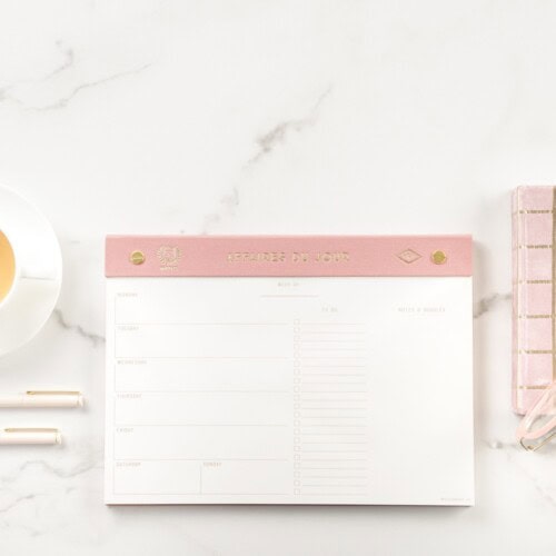 A weekly planner with a pink header lies on a white marble surface. Next to it are a cup of tea, two pens, and a pink notebook with gold accents from the happy journals, known for their elegant style and strict anti spam policy.