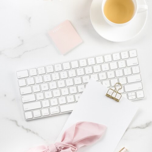A white keyboard, blank notepad with a gold clip, pink bow, sticky note, and a cup of tea on marble create a bright minimal workspace, perfect for organizing your thoughts or reviewing the happy journals policies.