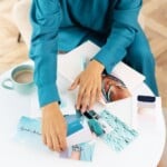 Person in a teal outfit arranging blue-toned photos and color swatches on a round white table with a cup of coffee, notebook, and pen—perhaps reflecting on the benefits of journal writing during their creative process.
