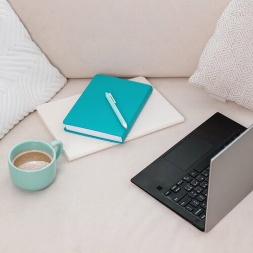 A cup of coffee, a closed laptop, a turquoise notebook with a pen for exploring the benefits of journal writing, and a white folder rest on a light couch with white pillows.