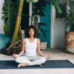 A woman sits cross-legged on a yoga mat outdoors, meditating with her eyes closed. Surrounded by lush potted plants and a turquoise wall, she reflects calmly, using journal prompts for forgiveness to guide her peaceful practice.