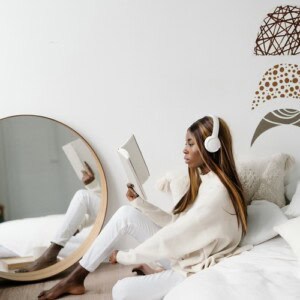 A woman in white clothes sits on a bed reading a book about mental health journaling prompts and wearing headphones. She is next to a round mirror on the floor, with decorative wall art above the bed on a white wall.