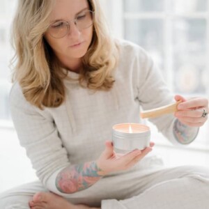 A woman with long blonde hair and glasses, dressed in light-colored loungewear, holds a lit candle and a piece of wood—her colorful tattoos on display. Sitting indoors, she finds calm as she works to rebuild confidence within herself.