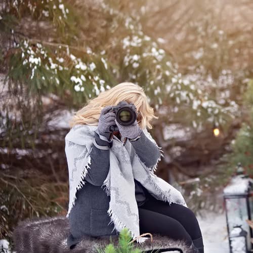 A person with long blonde hair, wearing a gray coat and gloves, sits outdoors in a snowy setting and takes a photo with a camera—capturing confidence boosters amid pine trees and snow in the background.