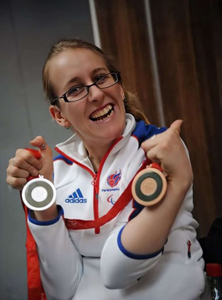 Fran whitaker, featured in the happy journals, smiles in a white and blue adidas sports jacket as she proudly holds up two medals. She wears glasses and has a paralympic logo on her jacket.