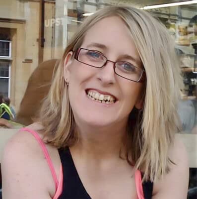 Fran Whitaker The Happy Journals 3 Fran whitaker of the happy journals, with blonde hair, glasses, and a black tank top with pink straps, smiles while sitting outside a café. The background shows a window with reflections and a person in a yellow vest.