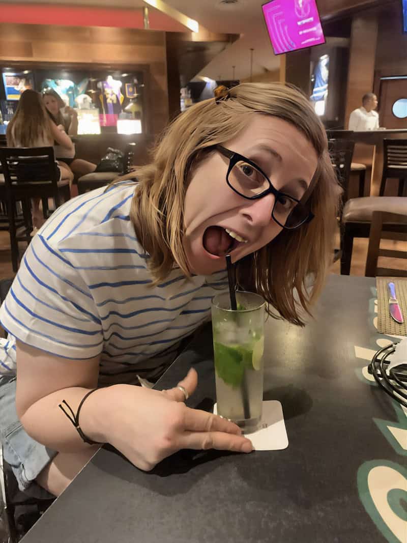 Fran whitaker of the happy journals playfully poses with her mouth open, wearing glasses and a striped shirt, next to a mojito on the table in a lively indoor setting. She points at the drink and smiles enthusiastically.