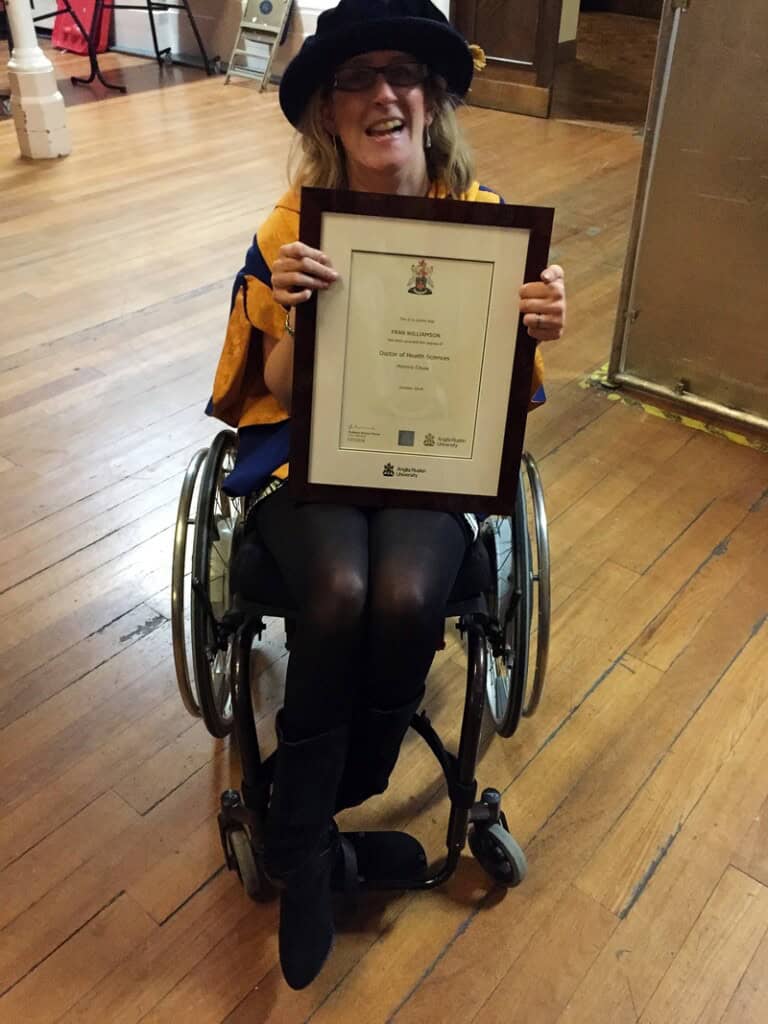 A smiling woman in a wheelchair, wearing a graduation cap and gown, holds a framed diploma while sitting on a wooden floor indoors—an inspiring fran whitaker - the happy journals moment.