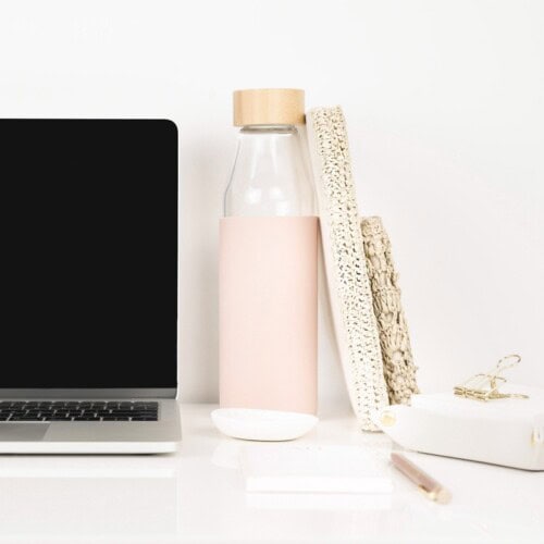 A laptop sits on a white desk next to a glass water bottle with a pink sleeve, two textured notebooks perfect for exploring different types of journaling, a gold paper clip holder, and a pen, all arranged neatly in a minimalist workspace.