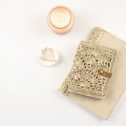 A beige woven pouch with a button rests on a folded tan cloth next to a lit candle in a pink holder and a small dish with cotton swabs, ideal for cozy evenings exploring different types of journaling, all set against a white background.