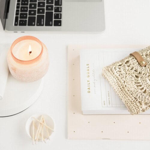 A laptop, a lit candle, a daily goals notepad for different types of journaling, a woven pouch, and a small dish with cotton swabs are arranged neatly on a white desk.