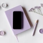 A lavender notebook for mindful journaling with a black smartphone on top, a matching pen, purple tealight candles, and a sprig of small purple flowers arranged on a white surface.