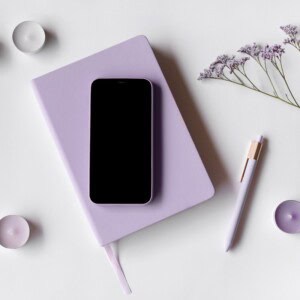 A lavender notebook for mindful journaling with a black smartphone on top, a matching pen, purple tealight candles, and a sprig of small purple flowers arranged on a white surface.