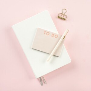 A white notebook, a to do notepad, a white pen, and a gold binder clip are arranged on a light pink background, inspiring ideas on how to create positive thoughts as you plan your day.