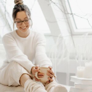 A smiling person wearing glasses, a white sweater, and cozy socks sits indoors by a bright window, holding a coffee mug—a peaceful self-care routine. White candles and decorative branches in the background create a warm, relaxed atmosphere.
