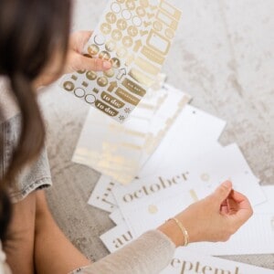 A woman in a cream jumper and grey jeans, adding gold monthly stickers to a journal as part of her monthly reflection practice