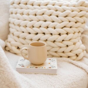 A cream knitted cushion on a cream sofa behind a book with a mug of coffee, ready for the user to try some of our easy self-care activities