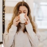 A person with long wavy hair wearing a light sweater is standing indoors, holding a white mug with both hands and drinking from it with eyes closed—enjoying a peaceful moment that highlights the benefits of self-care. The background is softly lit and slightly blurred.