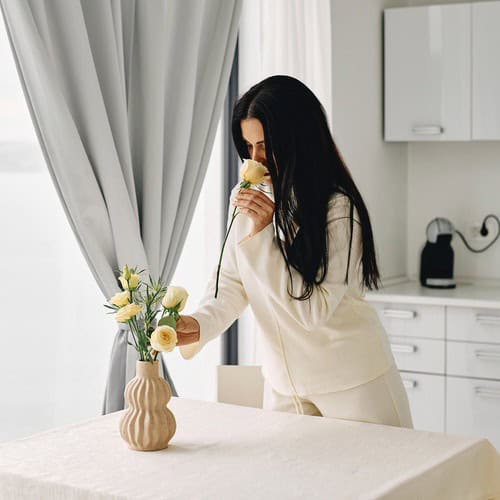 Morning journaling 2 A woman with long brown hair, dressed in a cream tracksuit, smelling some yellow roses as she sits down for some morning journaling