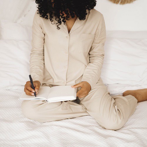 Morning journaling prompts 1 A woman with curly brown hair, dressed in a cream tracksuit, using some morning journaling prompts in her journal