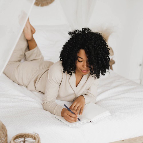Morning journaling prompts 3 A woman with curly brown hair, dressed in a cream tracksuit, using some morning journaling prompts in her journal