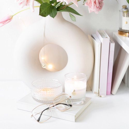 30 day guided journal 1 A white circular vase with pink flowers sits beside stacked books and a 30 day guided journal, a glass candle, a textured container, and eyeglasses on a white surface. Warm fairy lights add a soft glow.